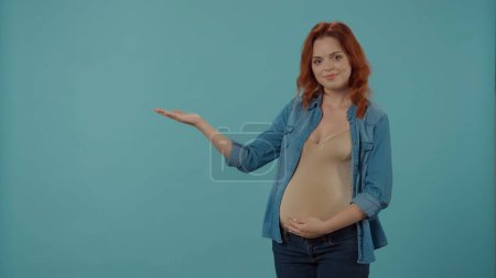 Téléchargez les photos : Une femme rousse, qui attend un bébé, tient un ventre enceinte et pointe avec sa paume vers un espace vide pour la publicité. Une femme enceinte en studio sur fond bleu - en image libre de droit