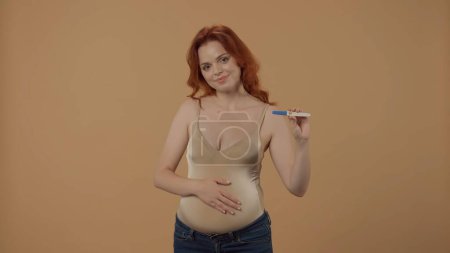 Téléchargez les photos : Une femme enceinte tient un test de grossesse avec un résultat positif dans un studio sur fond orange. Une femme heureuse tient son gros ventre avec sa main - en image libre de droit