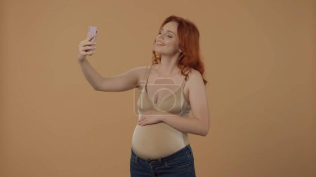 Foto de Feliz momento. Una mujer embarazada pelirroja toma una selfie, habla en una videollamada usando un teléfono inteligente. Mujer embarazada en el estudio sobre un fondo naranja. El concepto embarazo, maternidad - Imagen libre de derechos