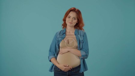 Foto de Retrato de una joven embarazada abrazando su gran vientre con las manos. Mujer embarazada aislada en azul. El concepto de embarazo, maternidad y amor - Imagen libre de derechos