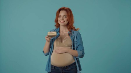 Téléchargez les photos : Une femme enceinte tient une assiette avec un morceau de gâteau. Femme en studio sur fond bleu. Envies de grossesse - en image libre de droit