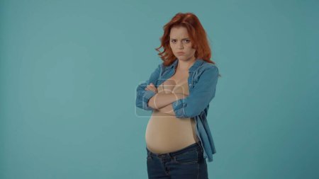Foto de Una mujer embarazada pelirroja se molesta, ofendida. Una mujer con los brazos cruzados en el pecho se para en el estudio sobre un fondo azul. Embarazo y emociones, humor cambiante, hormonas - Imagen libre de derechos