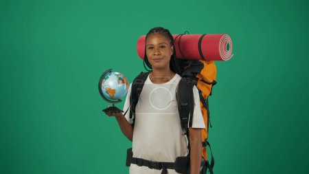 Téléchargez les photos : Une voyageuse tenant un globe et regardant la caméra. Portrait d'une touriste en tenue décontractée avec sac à dos. Voyager en temps libre concept. Isolé sur fond vert. - en image libre de droit