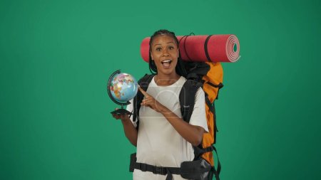 Téléchargez les photos : Une voyageuse tenant un globe et souriant à la caméra. Portrait d'une touriste en tenue décontractée avec sac à dos. Voyager en temps libre concept. Isolé sur fond vert. - en image libre de droit