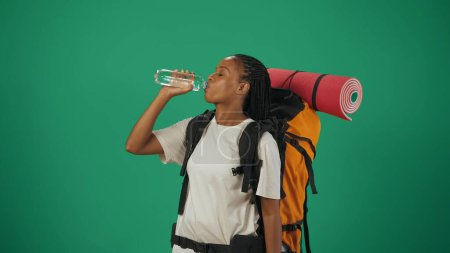 Téléchargez les photos : Une voyageuse boit de l'eau de la bouteille. Portrait d'une touriste en tenue décontractée avec sac à dos. Voyager en temps libre concept. Isolé sur fond vert. - en image libre de droit