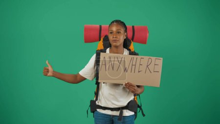 Photo for Woman hitchhiking, holding cardboard sign with word anywhere. Portrait of a female tourist in casual clothes with backpack. Travelling in spare time concept. Isolated on green background. - Royalty Free Image