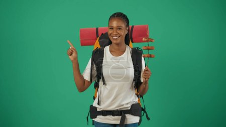 Photo for Woman traveller holding stick with sausages and pointing at the empty area. Portrait of a female tourist in casual clothes with backpack. Travelling in spare time concept. Isolated on green background - Royalty Free Image