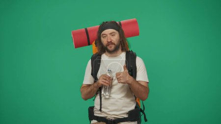 Photo for Man traveller holds bottle of water and shows thumbs up. Portrait of a male tourist in casual clothes with backpack. Travelling in spare time concept. Isolated on green background. - Royalty Free Image