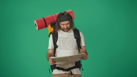 Photo for Man traveller holding paper map, looking clueless. Portrait of a male tourist in casual clothes with backpack. Travelling in spare time concept. Isolated on green background. - Royalty Free Image