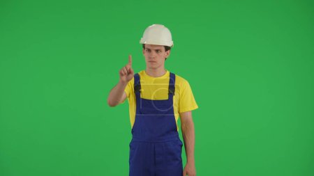 Téléchargez les photos : Écran vert moyen isolé chroma vidéo clé d'un jeune ouvrier de la construction glisser, tapant sur l'écran virtuel en face de lui. Construction, réparation, fabricant, publicité de l'entreprise - en image libre de droit