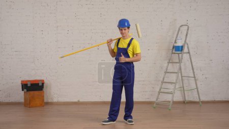 Photo for Full length shot of a smiling young worker standing in the room with a roller on his shoulder, looking at the camera and giving a thumbs up. Construction, paint, manufacturer, company advertisement. - Royalty Free Image