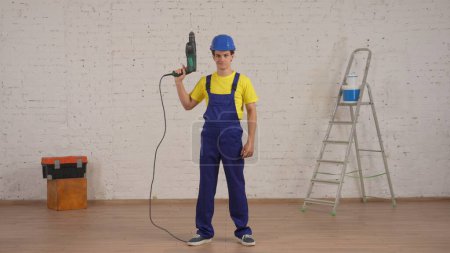 Photo for Full length shot of a smiling young worker standing in the room raising a drill, perforator in his hand up in the air and looking at the camera. Construction, paint, manufacturer, company - Royalty Free Image