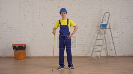 Photo for Full length shot of a young construction foreman standing in the room under renovation, leaning on the construction tape measure. Repairing, construction, company advertisement. - Royalty Free Image