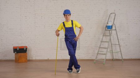 Photo for Full length shot of a young construction foreman standing in the room under renovation, leaning on the construction tape measure. Repairing, construction, company advertisement. - Royalty Free Image