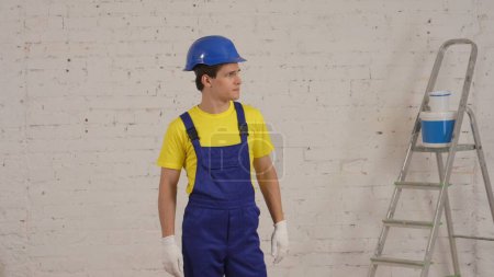 Téléchargez les photos : Plan moyen d'un jeune contremaître de chantier debout dans la pièce en rénovation, portant uniforme et gants, regardant sur le côté. Réparation, publicité de l'entreprise. - en image libre de droit