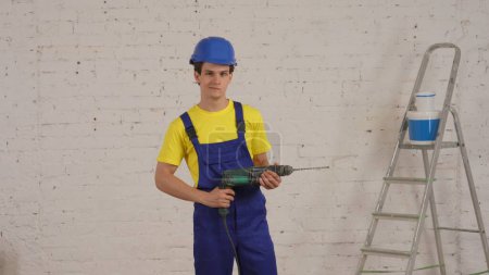 Photo for Medium shot of a smiling young worker standing in the room holding a drill, perforator in his hand like a gun and looking at the camera. Construction, repair, manufacturer, company advertisement. - Royalty Free Image
