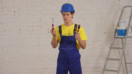 Photo for Medium shot of a confused young construction worker standing in the room under renovation, deciding which screwdriver, turn-screw to use. Repairing, manufacturer, company advertisement. - Royalty Free Image