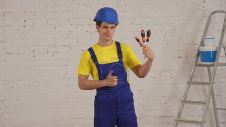 Photo for Medium shot of a young construction worker standing in the room under renovation, holding screwdrivers, turn-screws, giving thumbs up, recommending the product. Repairing, manufacturer, company - Royalty Free Image