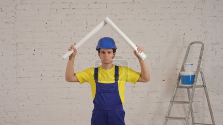 Téléchargez les photos : Plan moyen d'une vidéo souriante d'un jeune ouvrier de la construction levant deux plans enroulés au-dessus de sa tête, imitant un toit. Construction, fabricant, publicité de l'entreprise. Sécurité, fiabilité. - en image libre de droit