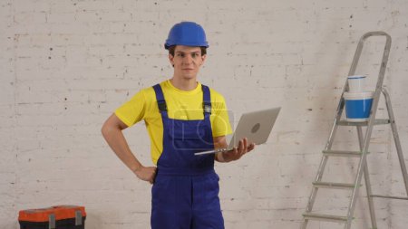Photo for Medium shot of a young construction worker standing in the room, holding a laptop, researching, working, smiling at the camera. Construction, repairing, manufacturer, company advertisement. - Royalty Free Image