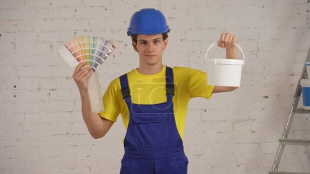 Photo for Medium shot of a young construction worker standing in the room under renovation, showing a bucket full of paint for walls and a palette. Construction, paint, manufacturer, company advertisement. - Royalty Free Image