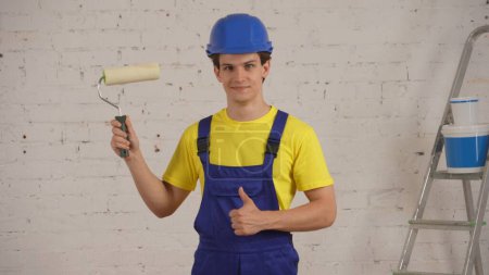 Photo for Medium shot of a young construction worker standing in the room under renovation with a roller in his hand, giving a thumbs up, recommending the product. Repairing, product, company advertisement. - Royalty Free Image