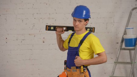 Photo for Medium shot of a smiling young construction worker wearing a tool belt, holding a construction water level on his shoulder, smiling and looking at the camera confidently. Repair, company advertisement - Royalty Free Image
