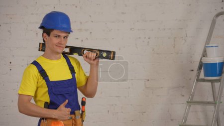Photo for Medium shot of a smiling young construction worker wearing a tool belt, holding a construction water level on his shoulder, smiling and giving a thumbs up. Repair, company advertisement. - Royalty Free Image