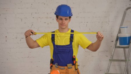 Téléchargez les photos : Plan moyen d'un jeune ouvrier de la construction souriant portant une ceinture à outils, dépliant le ruban à mesurer de construction devant la caméra. Construction, réparation, fabricant, publicité de l'entreprise. - en image libre de droit
