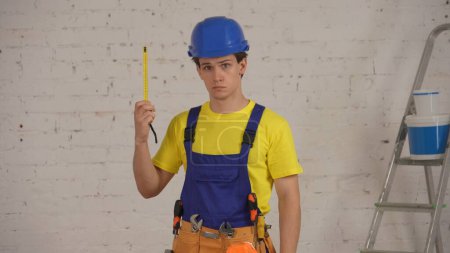 Téléchargez les photos : Plan moyen d'un jeune ouvrier de la construction souriant portant une ceinture à outils, tenant un ruban de construction déplié et regardant la caméra. Construction, réparation, fabricant, entreprise - en image libre de droit
