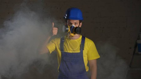 Photo for Medium shot of a construction worker standing in the room filled with smoke, happy to wear a respirator and giving a thumbs up. Safety precautions, protection, manufacturer, company advertisement. - Royalty Free Image