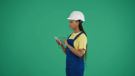 Foto de Captura de clave de croma aislada de pantalla verde media de una joven trabajadora de la construcción de piel oscura sosteniendo un teléfono inteligente y escribiendo, insertando datos mientras mira a su alrededor. Anuncio. Aplicación, sitio. - Imagen libre de derechos