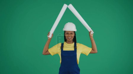 Photo for Medium green screen isolated chroma key shot of a dark-skinned young female construction worker raising two rolled up plans above her head, imitating a roof. Safety, reliability concept. - Royalty Free Image
