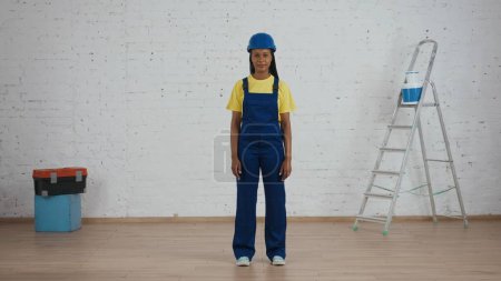 Photo for Full length shot of a smiling dark-skinned young female construction worker in a uniform and hardhat standing in the room under renovation. Manufacturer, company advertisement. - Royalty Free Image