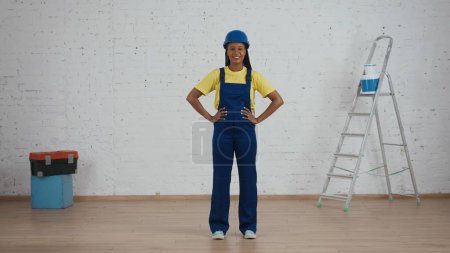 Photo for Full length shot of a smiling dark-skinned young female construction worker in a uniform and hardhat standing in the room under renovation with her hands on her hips. Manufacturer, company - Royalty Free Image