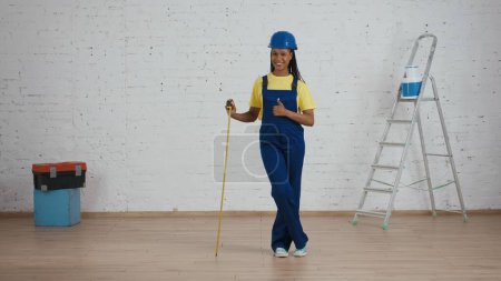 Photo for Full length shot of a dark-skinned young female construction foreman standing in the room under renovation, leaning on the construction tape measure and giving a thumbs up. Repairing, advertisement. - Royalty Free Image