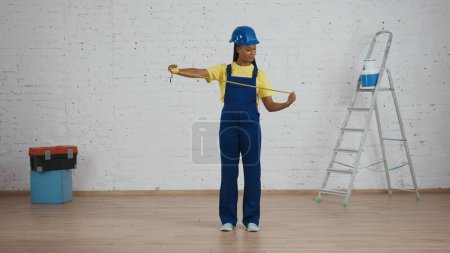 Photo for Full length shot of a dark-skinned young female construction foreman standing in the room under renovation, unfolding the construction tape measure. Repairing, company advertisement. - Royalty Free Image