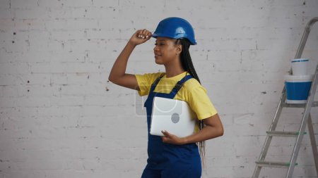 Photo for Medium shot of a dark-skinned young female construction worker standing in the room, holding a laptop under her arm, fixing a hardhat, turned to the side. Site, online, company advertisement. - Royalty Free Image