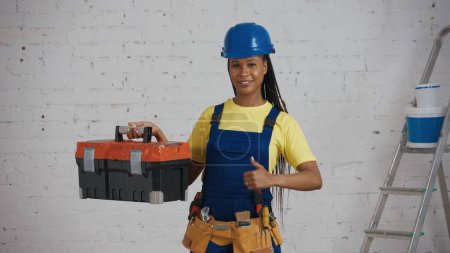 Photo for Medium shot of a dark-skinned young female construction worker standing in the room, wearing a tool belt, showing a tool box and giving a thumbs-up. Construction, repair, company advertisement. - Royalty Free Image