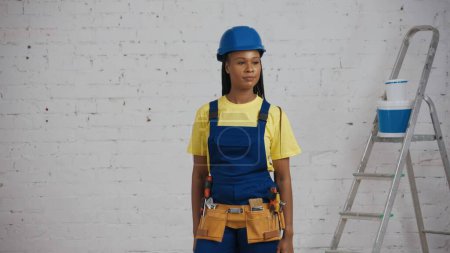 Foto de Foto media de una joven trabajadora de la construcción de piel oscura sonriente con un uniforme y un sombrero de trabajo de pie en la habitación, usando un cinturón de herramientas, mirando a su alrededor. Fabricante, anuncio de la empresa. - Imagen libre de derechos