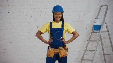 Photo for Medium shot of a smiling dark-skinned young female construction worker in a uniform standing in the room under renovation, loking at the camera with her hands on her hips. Company advertisement. - Royalty Free Image