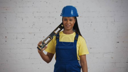 Photo for Medium shot of a dark-skinned young female construction worker standing in the room under renovation, holding a construction water level on her shoulder and smiling. Advertisement. - Royalty Free Image