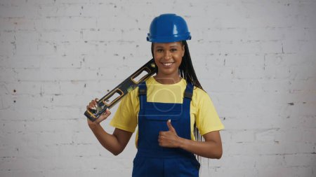 Photo for Medium shot of a dark-skinned young female construction worker standing in the room under renovation, holding a construction water level on her shoulder and giving a thumbs up. Advertisement. - Royalty Free Image