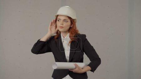 Téléchargez les photos : Femme architecte rousse penchant sa main à son oreille en essayant d'entendre, écoutant attentivement. Portrait d'une femme en casque dur avec des plans à la main dans le studio sur un fond gris - en image libre de droit