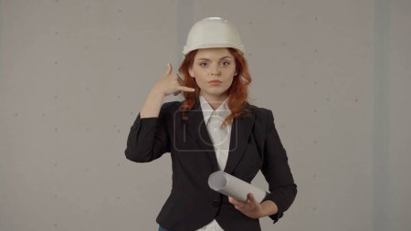 Photo for A woman architect makes a phone gesture near her head. Portrait of a woman in a hard hat with blueprints in her hand in the studio on a gray background - Royalty Free Image