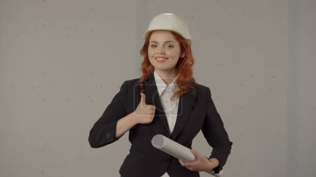 Foto de Una arquitecta muestra un pulgar hacia arriba. Retrato de una mujer en un casco con planos en la mano en el estudio sobre un fondo gris. Aceptación de obra terminada, objeto - Imagen libre de derechos