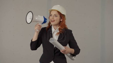Foto de Una mujer con un casco protector y dibujos en la mano habla en un megáfono. Una arquitecta corrige el trabajo usando un megáfono en un estudio sobre un fondo gris - Imagen libre de derechos