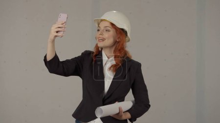 Photo for Woman architect talking on video call, taking selfie using smart phone. A woman in a helmet and with blueprints in a studio on a gray background - Royalty Free Image