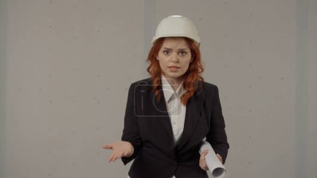 Photo for Angered woman speaks emotionally, gesticulating with her hands. Portrait of a woman in a protective helmet and with blueprints in the studio on a gray background - Royalty Free Image