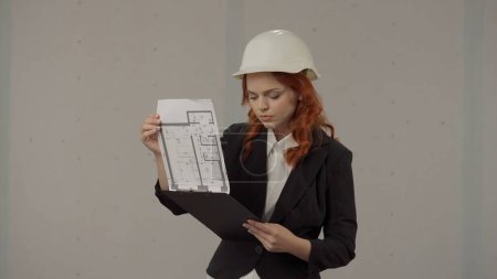 Foto de Una arquitecta estudia documentos, un plan de proyecto en un portapapeles. Retrato de una mujer con un casco protector, un traje de negocios en el estudio sobre un fondo gris - Imagen libre de derechos